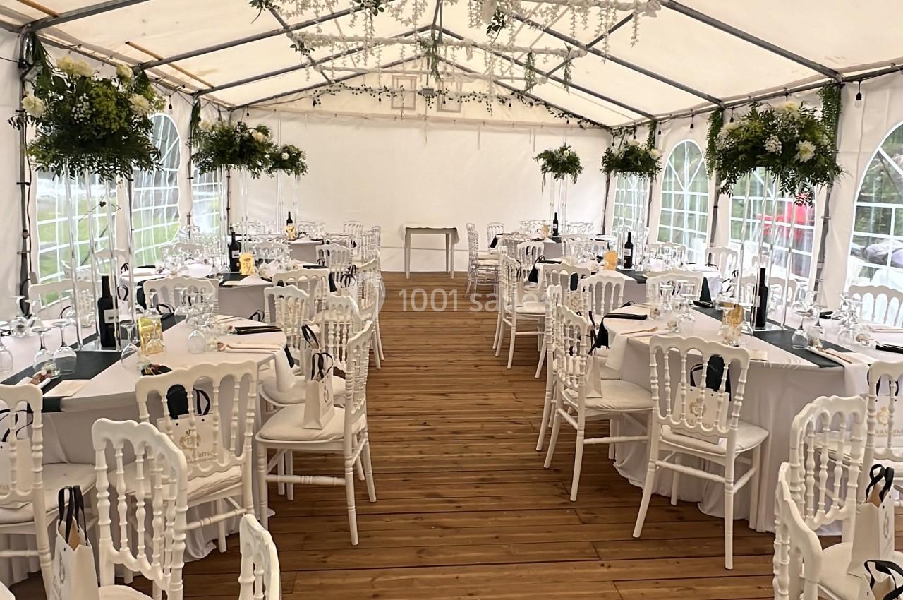 Salle de réception sous une tente blanche avec tables dressées, chaises blanches et décorations florales suspendues.