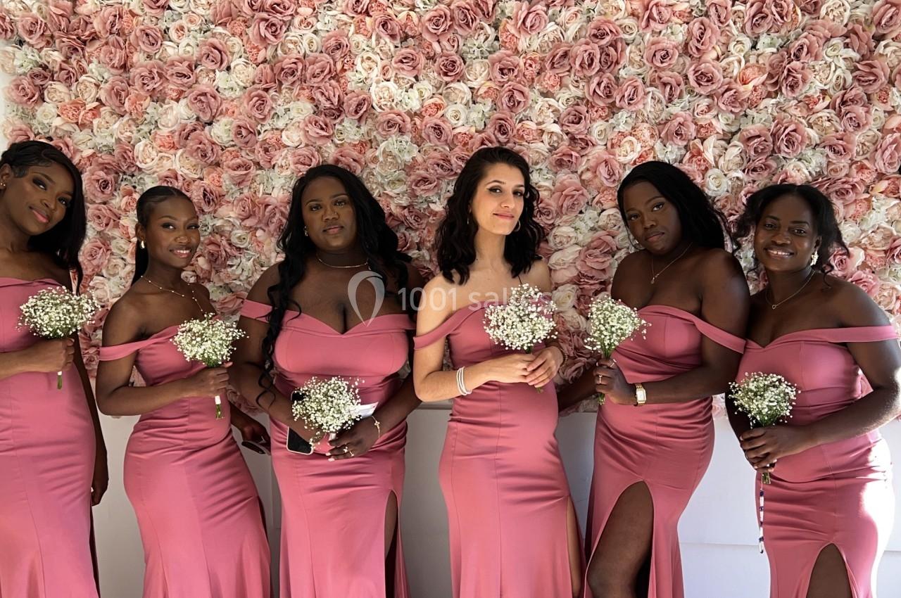 Six femmes en robes roses posent devant un mur couvert de roses, tenant des bouquets de fleurs blanches.