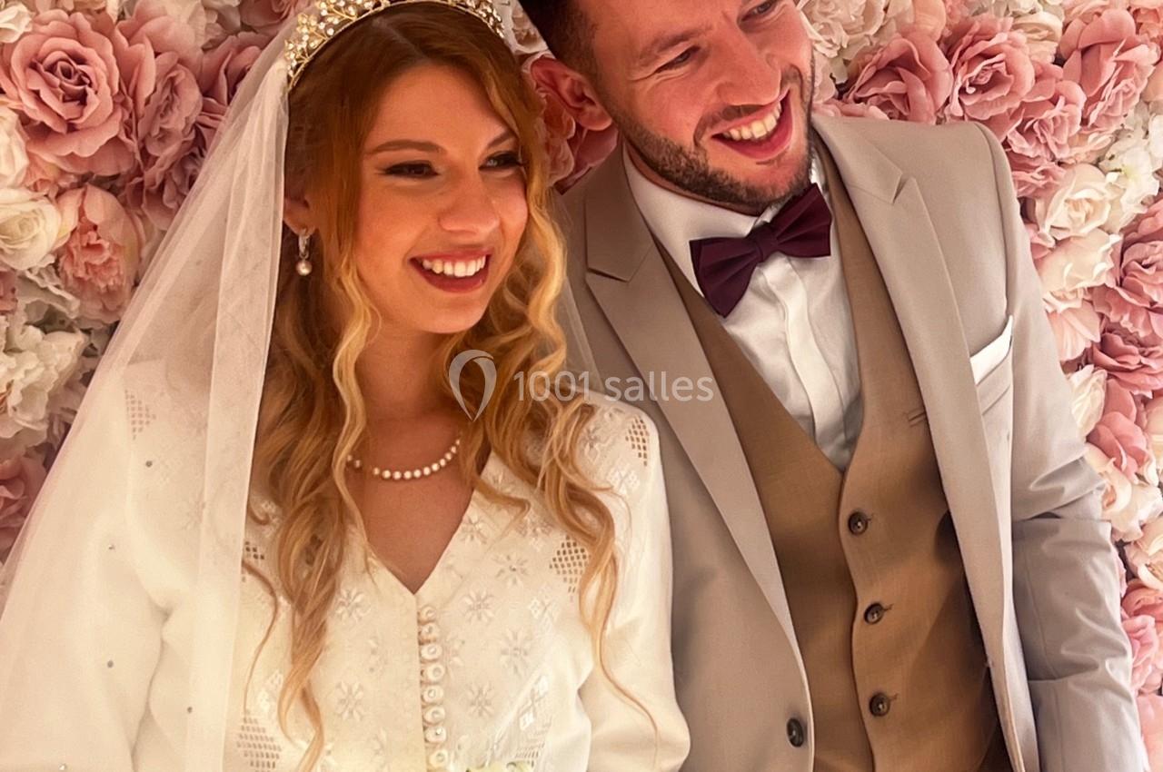 Un couple souriant en tenue de mariage pose devant un mur décoré de roses roses et blanches.