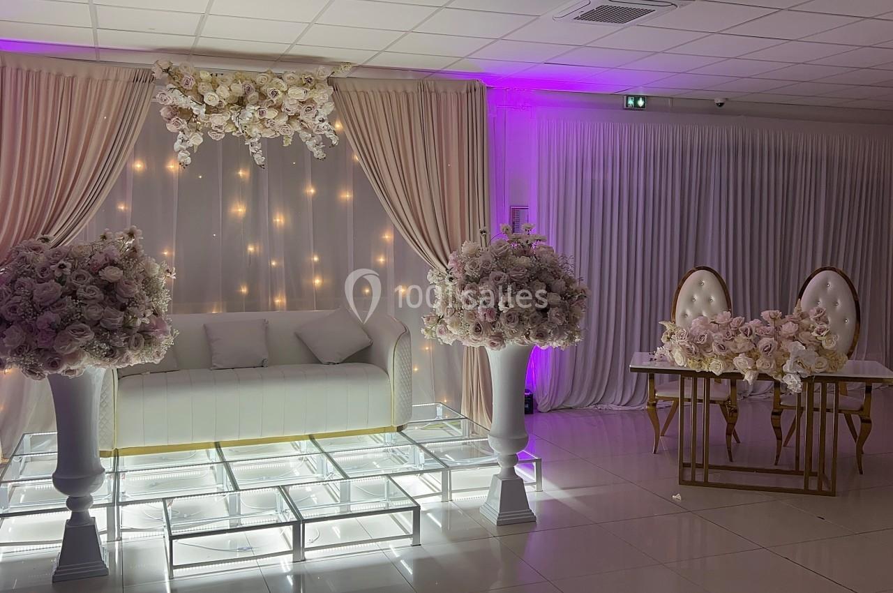 Décor de mariage avec canapé blanc, rideaux beiges, guirlandes lumineuses, fleurs blanches et table ornée de chaises dorées.