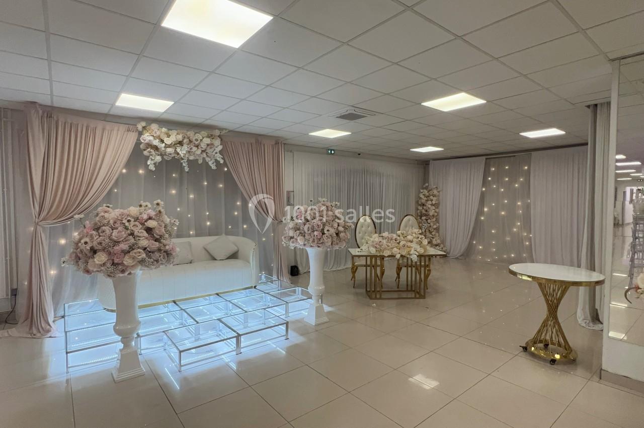 Salle décorée pour un événement avec rideaux roses, fleurs, canapé blanc, table en verre et chaises dorées.
