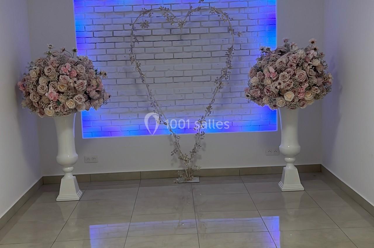 Deux bouquets de fleurs roses sur des colonnes blanches encadrent un cœur lumineux sur un mur en briques blanches.