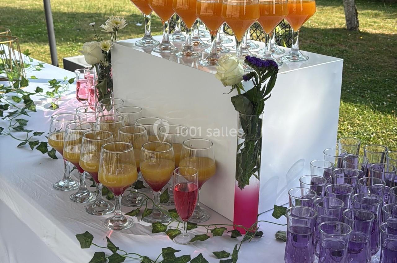 Table dressée avec des verres de cocktails colorés, décorée de feuillage, de roses blanches et violettes, en extérieur.