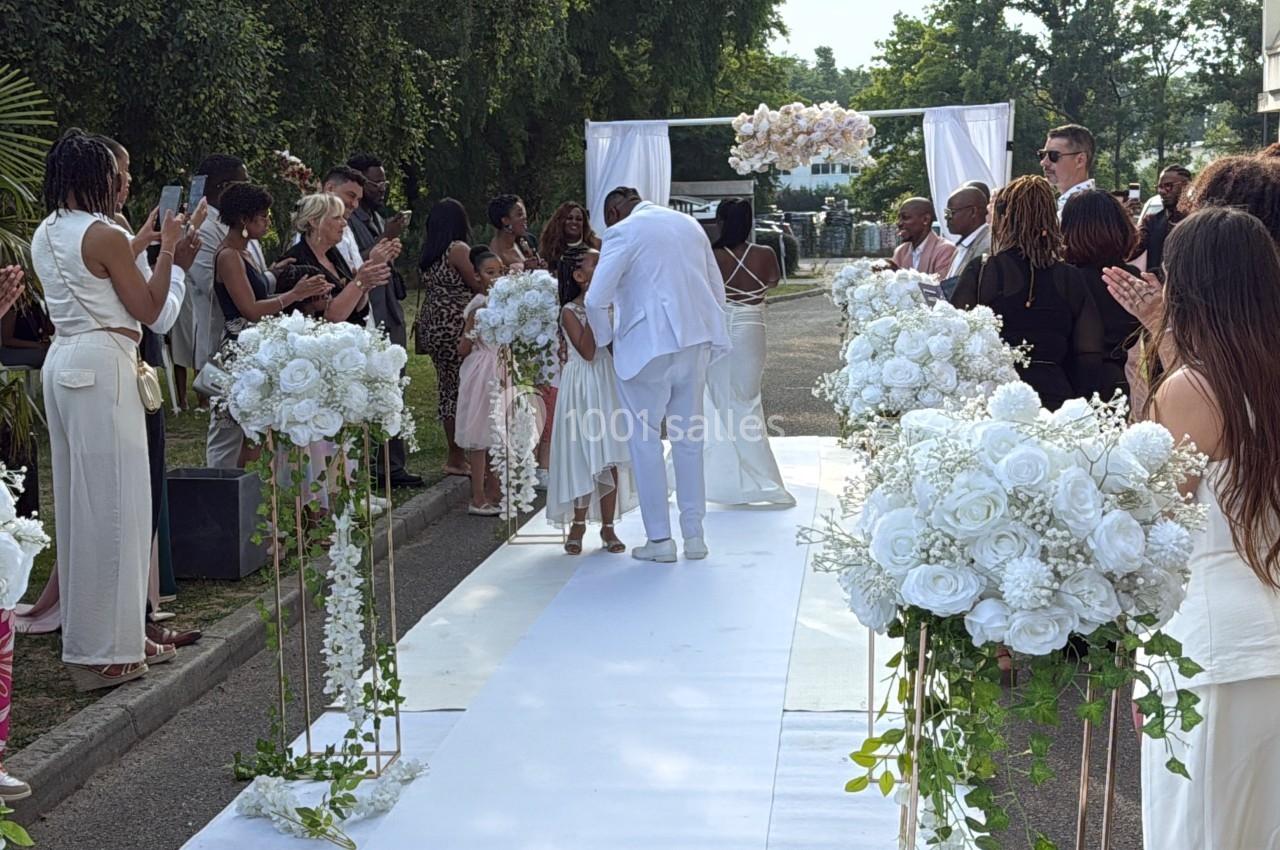 Un couple s'embrasse sur une allée décorée de fleurs blanches, entouré d'invités applaudissant lors d'une cérémonie en…