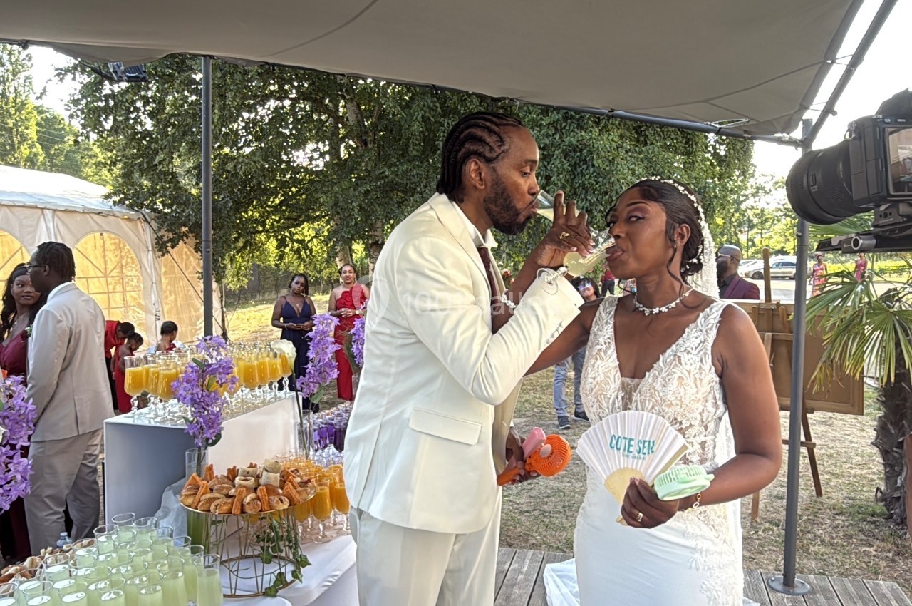 Un couple en tenue de mariage partage un moment complice lors d'une réception en plein air avec des invités en arrière-plan.