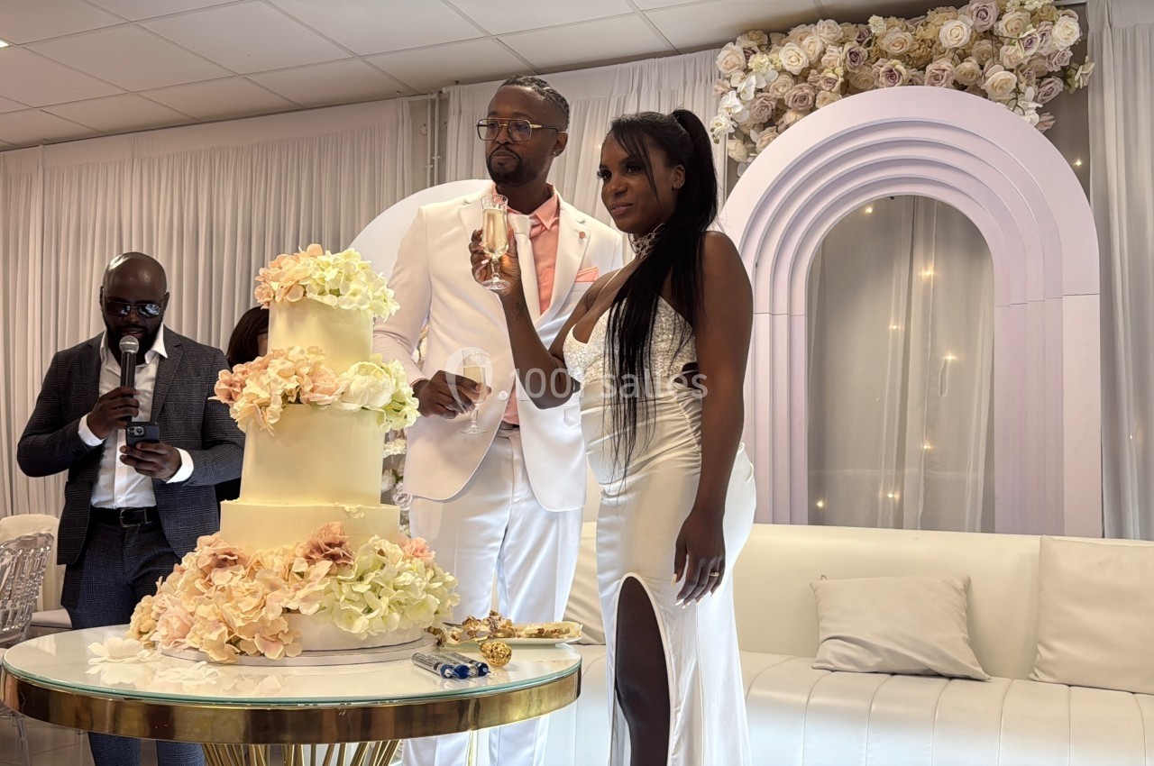 Un couple en tenue de mariage pose devant un gâteau décoré de fleurs, entouré d'invités dans une salle élégante.