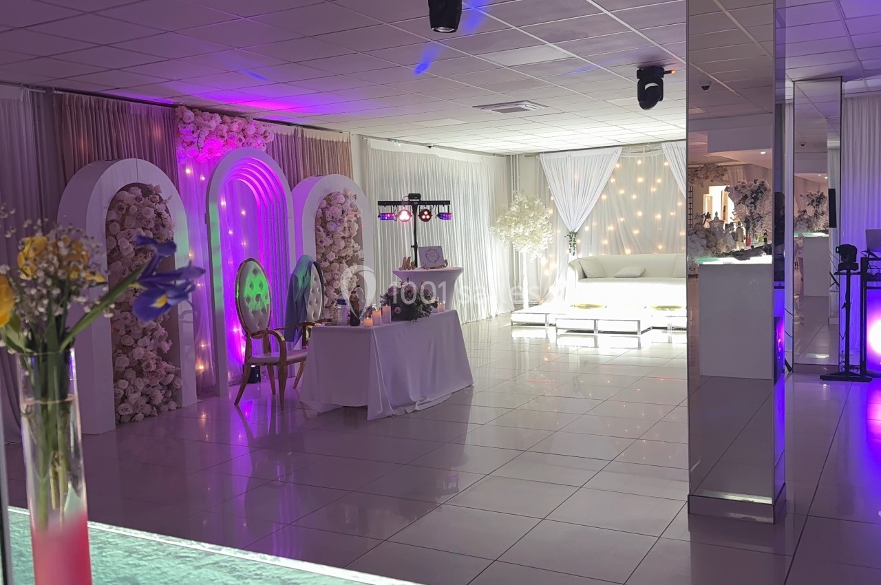 Salle décorée pour un événement avec des arches fleuries, éclairages colorés et mobilier blanc élégant.