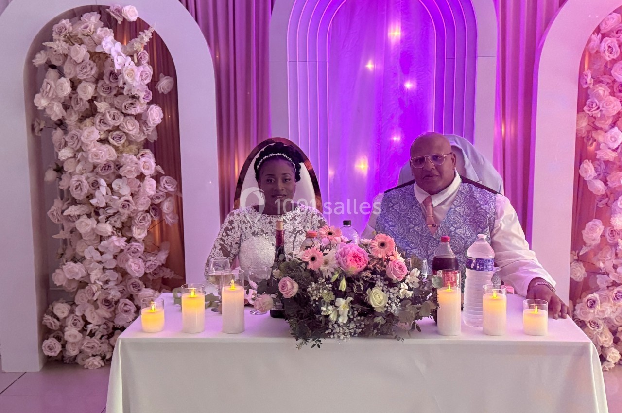 Un couple assis à une table décorée de fleurs et de bougies, dans un cadre festif avec des arches lumineuses.