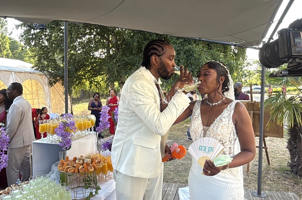 Un couple en tenue de mariage partage une boisson lors d'une réception en extérieur, entouré d'invités et d'un buffet.
