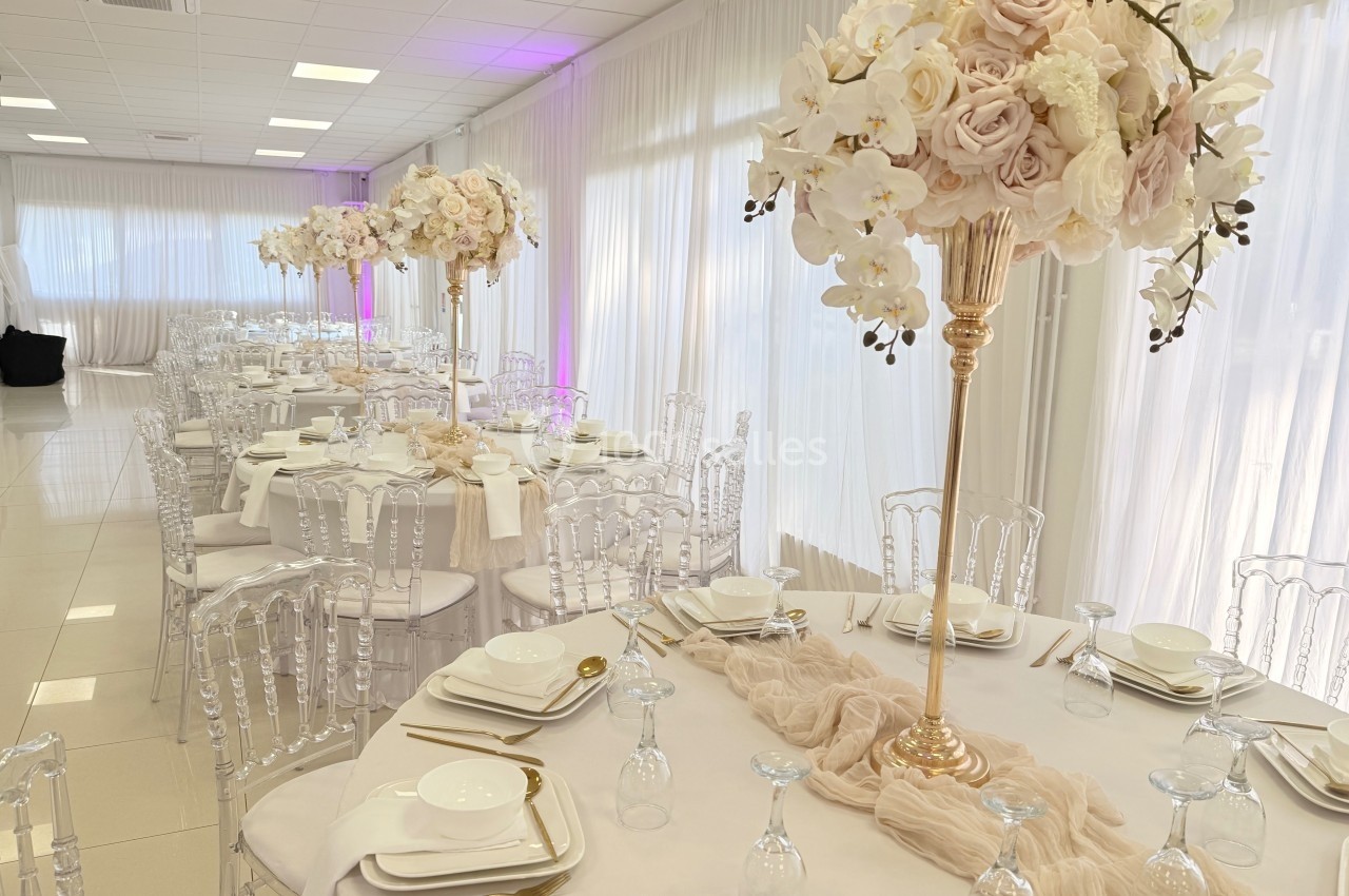 Salle de réception décorée avec des tables rondes, nappes blanches, vaisselle élégante et centres de table fleuris.