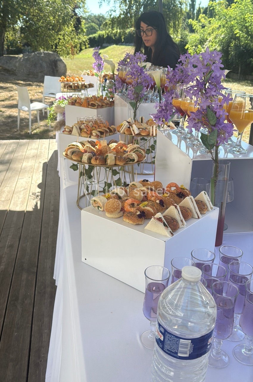 Buffet en plein air avec assortiments de sandwichs, viennoiseries, boissons et décorations florales violettes.