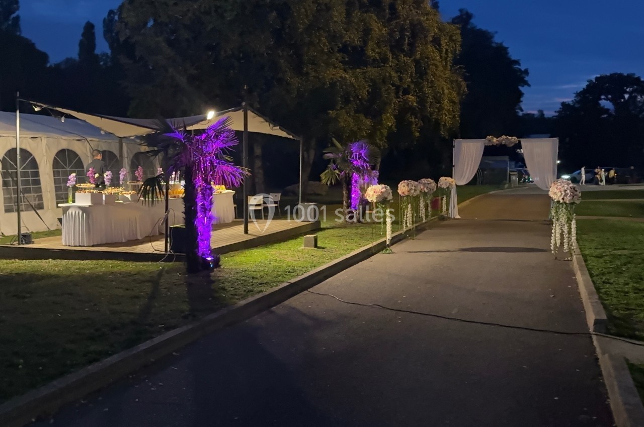 Allée décorée de fleurs et éclairages violets menant à une arche, avec une tente éclairée en arrière-plan, de nuit.