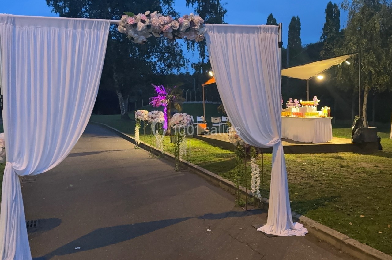 Arche décorée de rideaux blancs et de fleurs, installée dans un parc en soirée avec un stand éclairé en arrière-plan.