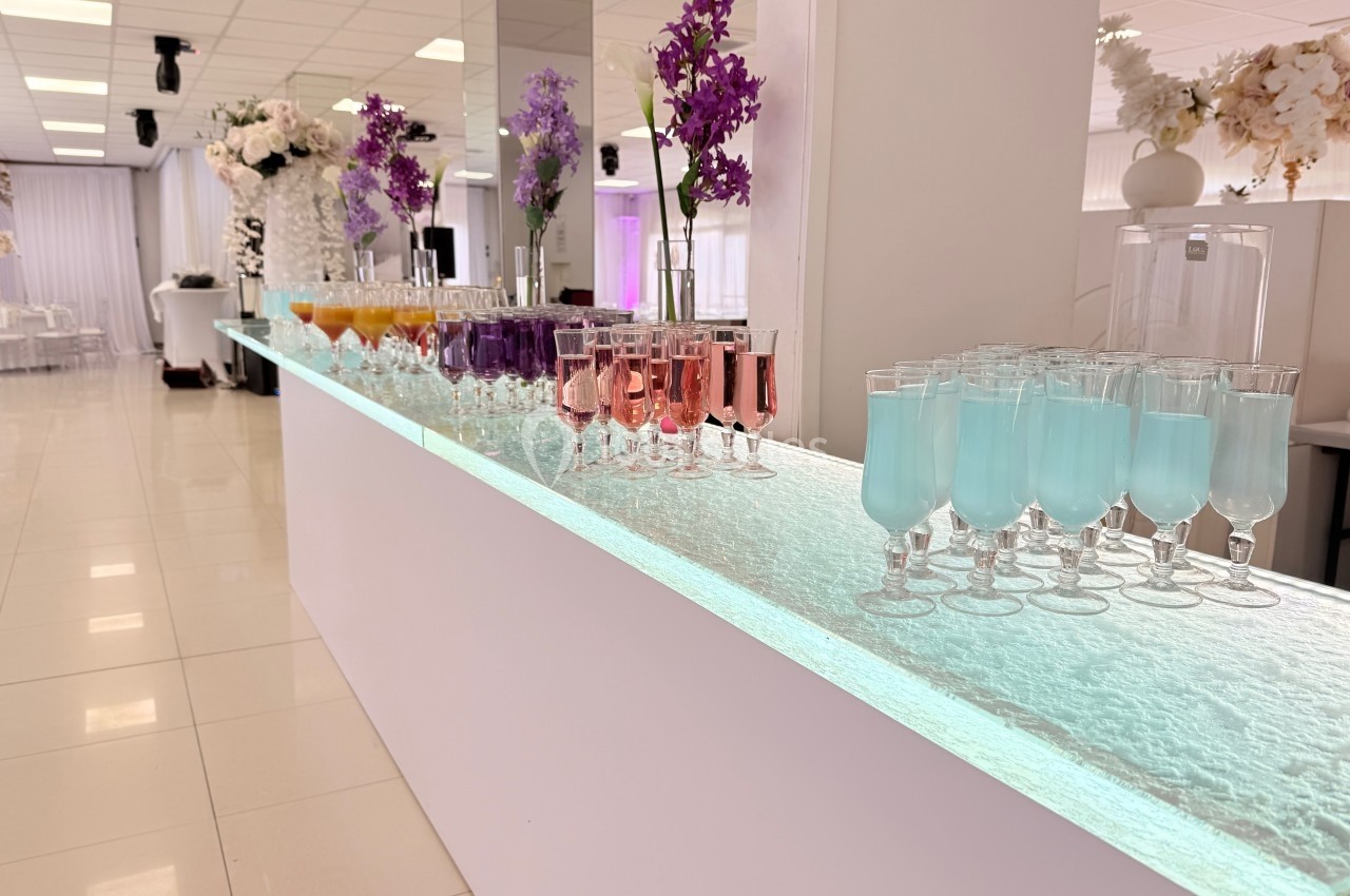 Verres de boissons colorées alignés sur un long comptoir lumineux dans une salle de réception décorée de fleurs.