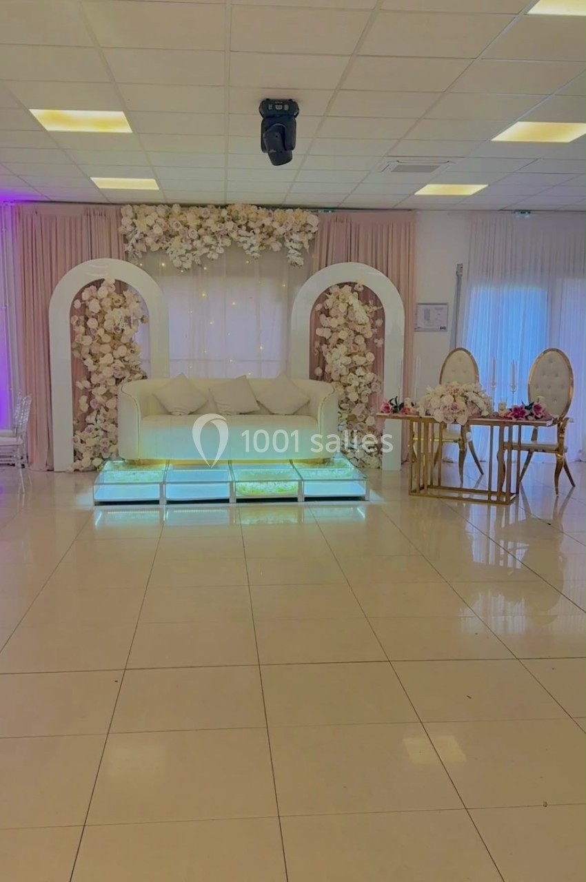 Salle de réception décorée avec un canapé blanc, des arches fleuries et une table élégante sur un sol carrelé.