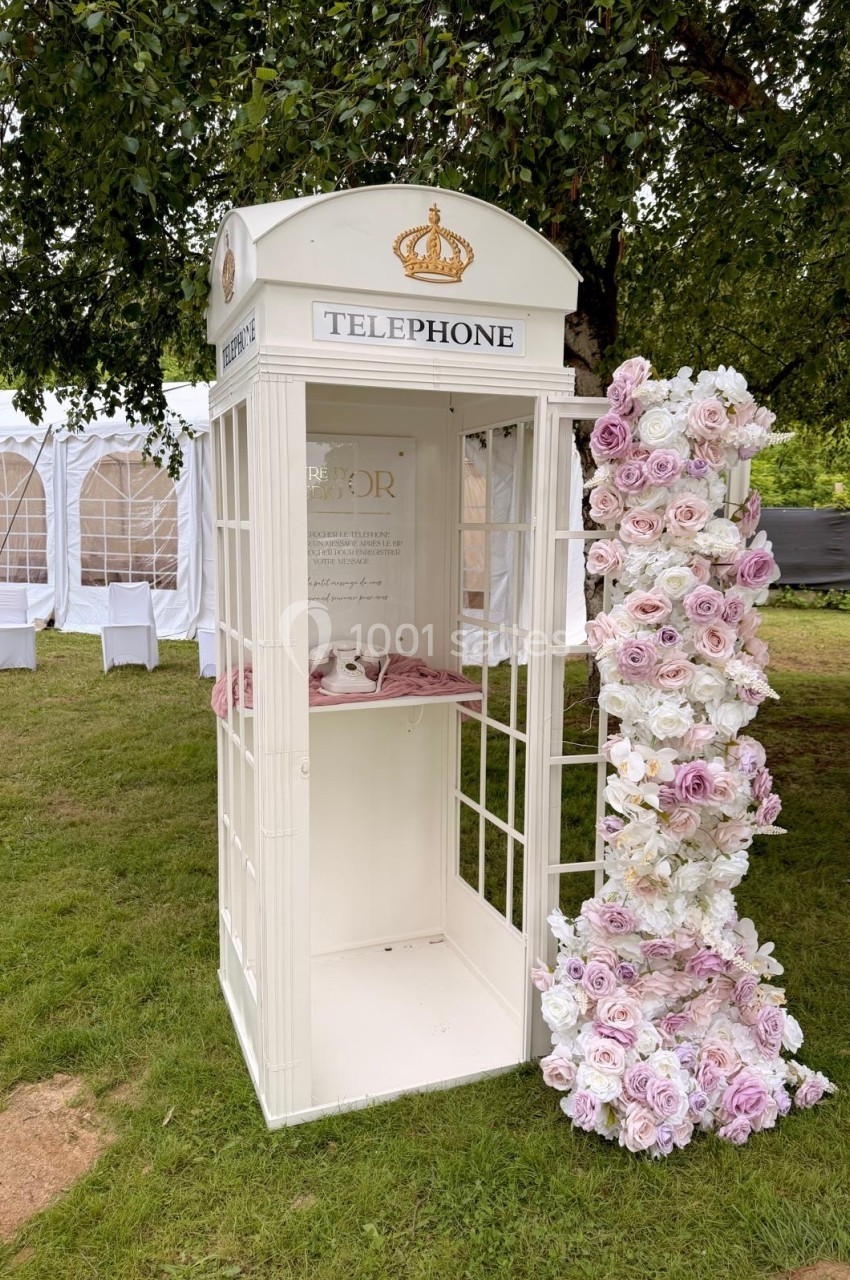 Cabine téléphonique blanche décorée de fleurs roses et blanches, installée sur une pelouse près de tentes blanches.