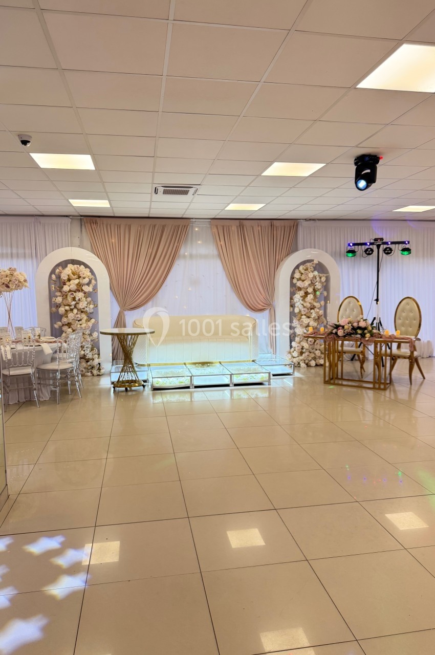 Salle de réception décorée avec des rideaux beiges, des arches fleuries blanches et un éclairage discret.