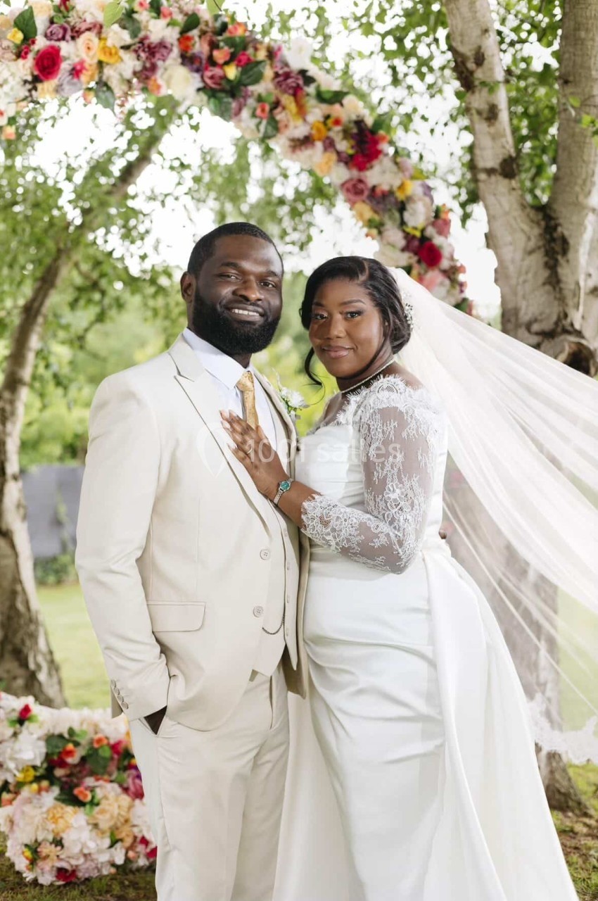 Un couple en tenue de mariage pose devant une arche fleurie dans un jardin verdoyant.