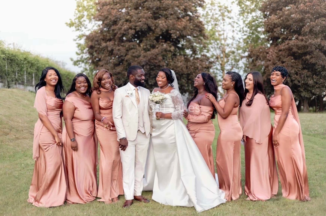 Un couple de mariés souriant entouré de demoiselles d'honneur en robes roses dans un jardin verdoyant.