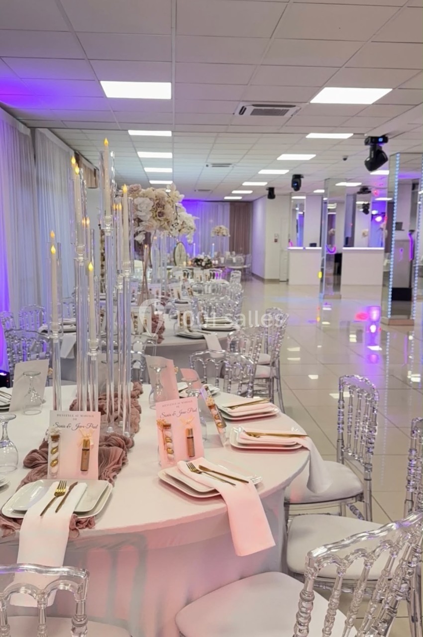 Salle de réception élégante avec tables rondes décorées de nappes roses, chandeliers et chaises transparentes.