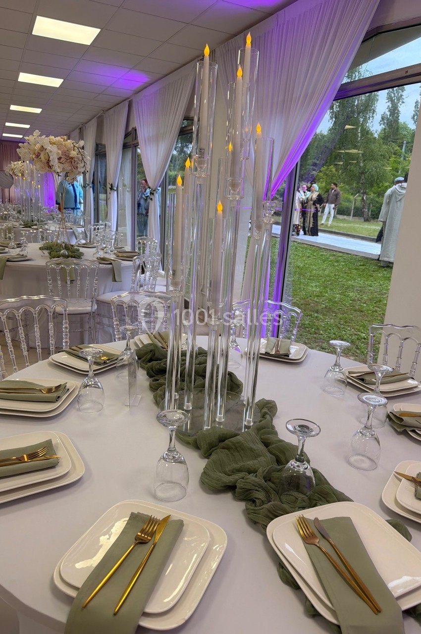 Salle de réception décorée avec des tables rondes, nappes blanches, couverts dorés et centre de table lumineux.