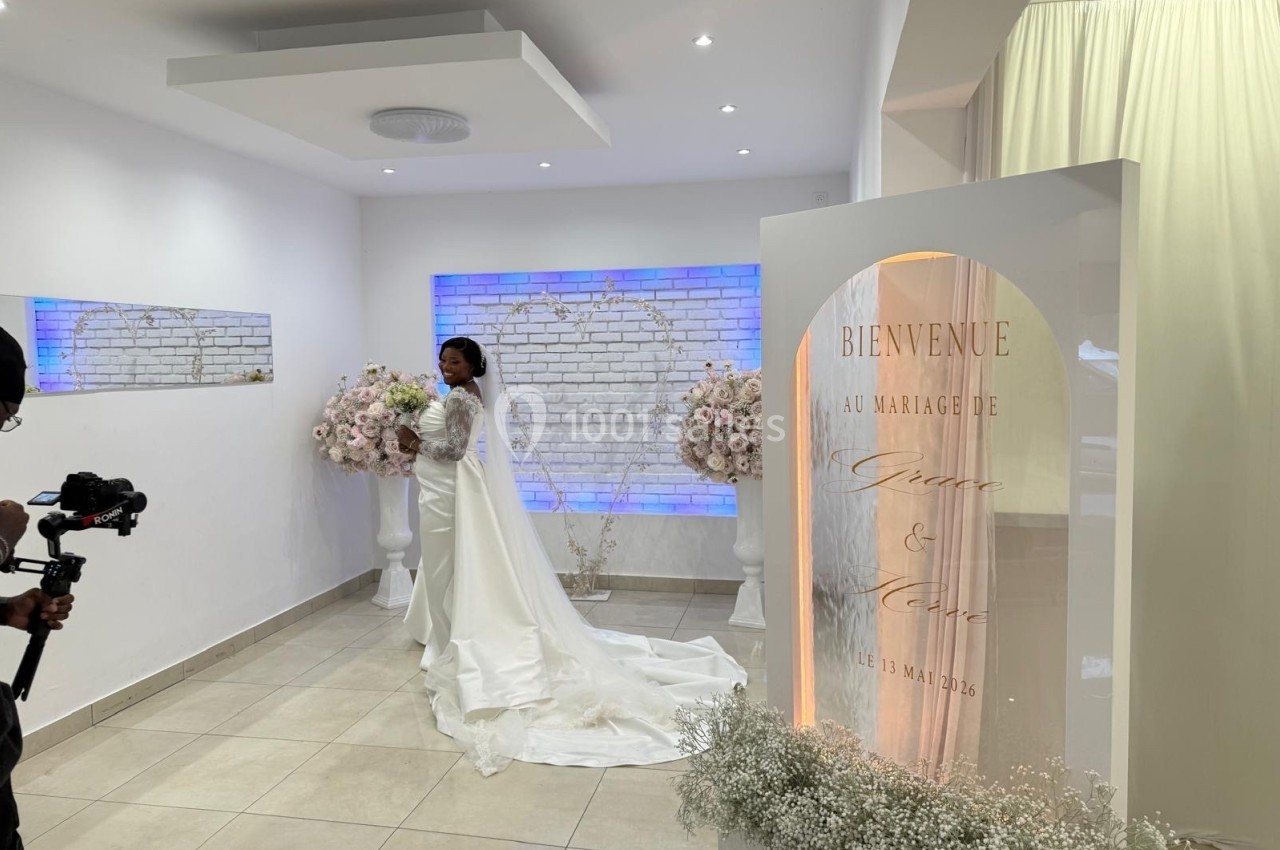 Une mariée en robe blanche pose devant une décoration florale et un panneau de bienvenue dans une salle lumineuse.