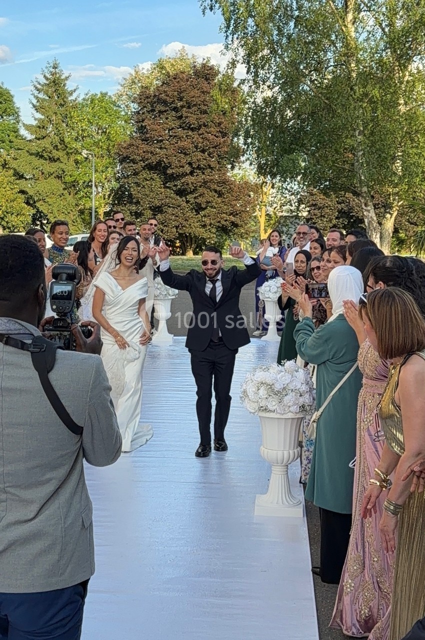 Un couple marche sur une allée blanche entouré d'invités applaudissant lors d'une cérémonie en plein air.