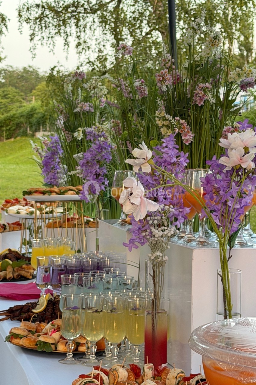 Buffet en plein air avec boissons colorées, brochettes, pâtisseries et décorations florales violettes et blanches.