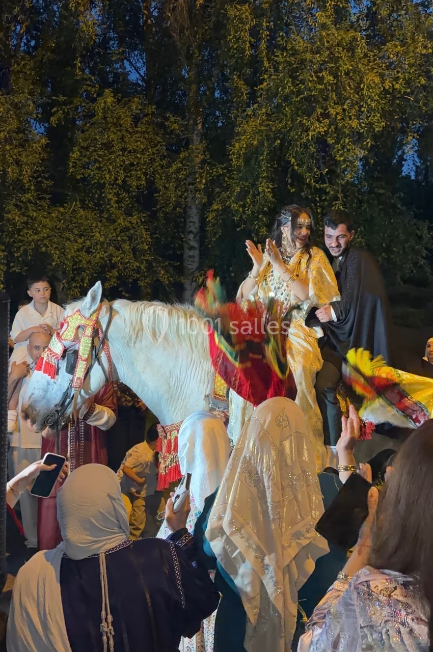 Un couple en tenue traditionnelle célèbre un événement sur un cheval blanc décoré, entouré de personnes festives.