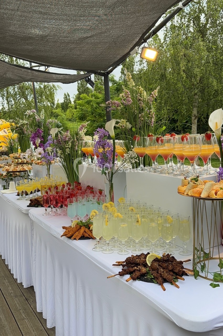 Buffet en plein air avec cocktails colorés, brochettes, fleurs décoratives et desserts sur des tables drapées de blanc.