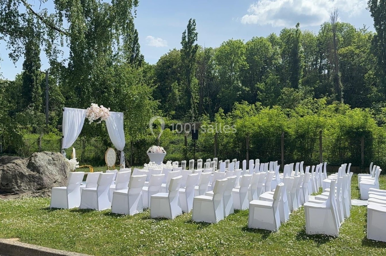 Chaises blanches alignées en extérieur, face à une arche décorée de fleurs, dans un cadre verdoyant.