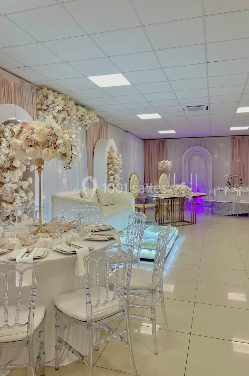 Salle de réception décorée avec des fleurs blanches et roses, tables dressées et chaises transparentes.