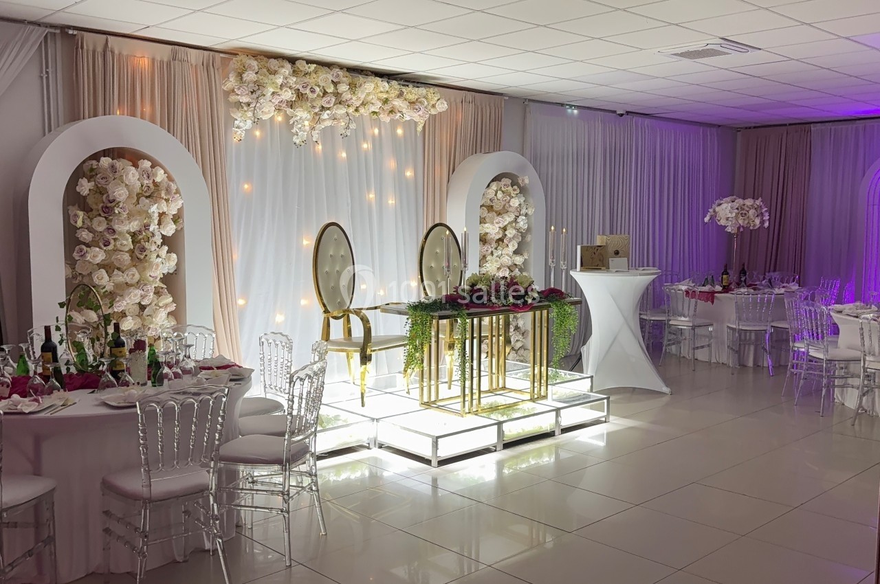 Salle de réception décorée avec des fleurs blanches, des chaises transparentes et une table d'honneur illuminée.