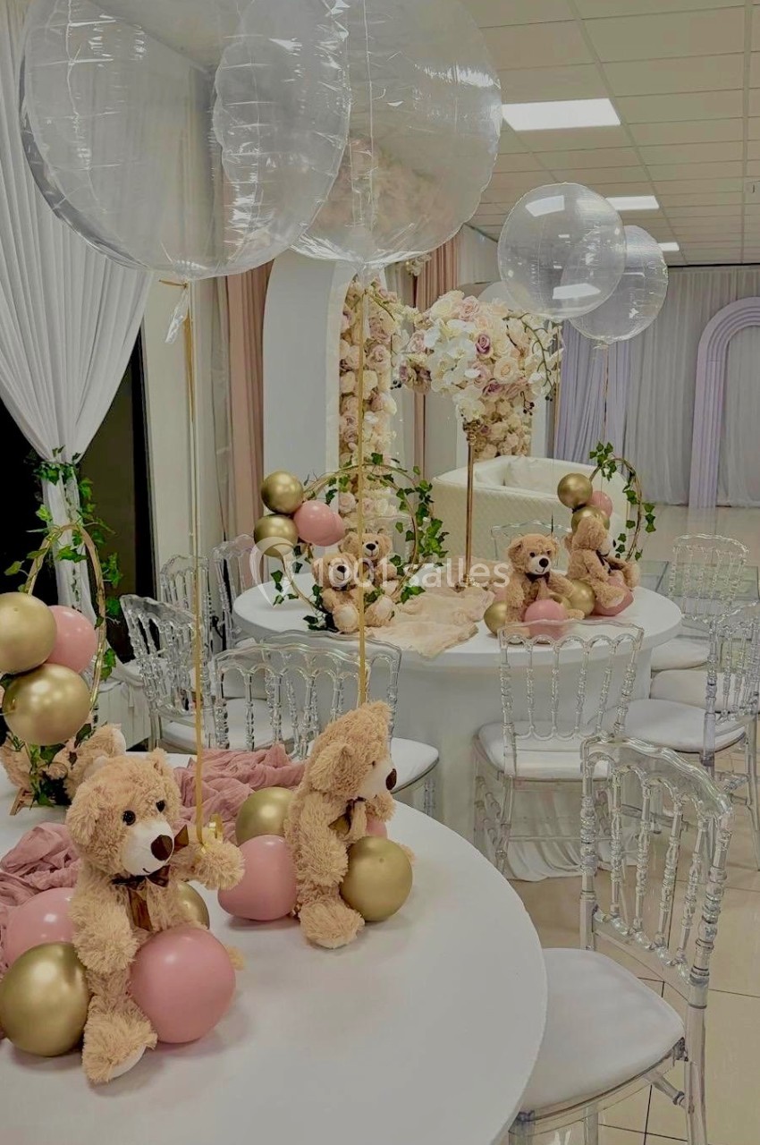 Décoration de table avec ours en peluche, ballons roses et dorés, et arches florales dans une salle lumineuse.