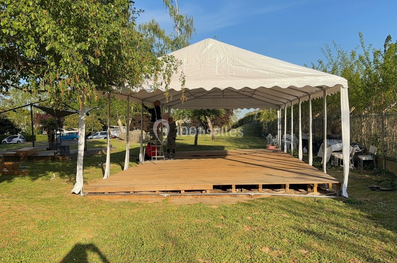 Tente blanche montée sur une structure en bois dans un espace extérieur verdoyant par temps ensoleillé.