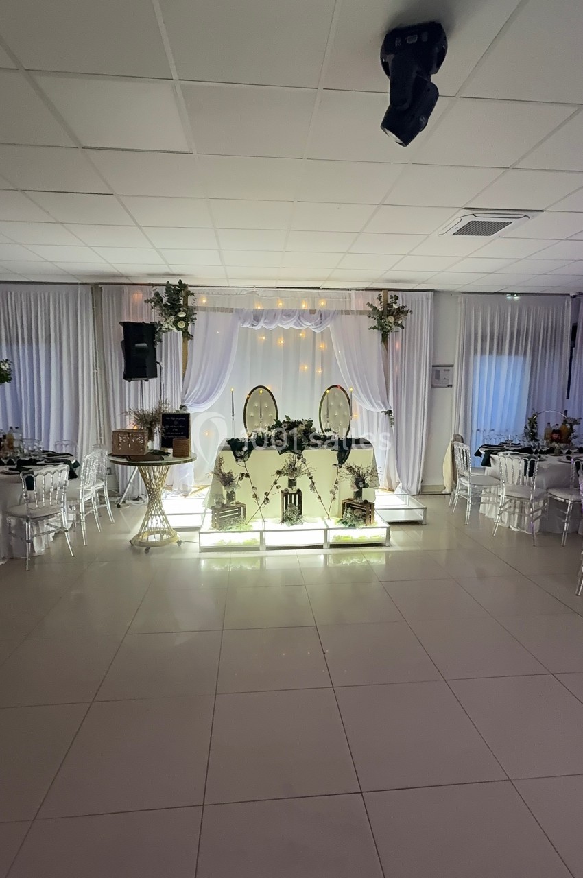 Salle de réception décorée avec une table d'honneur illuminée, ornée de fleurs et entourée de tables dressées.