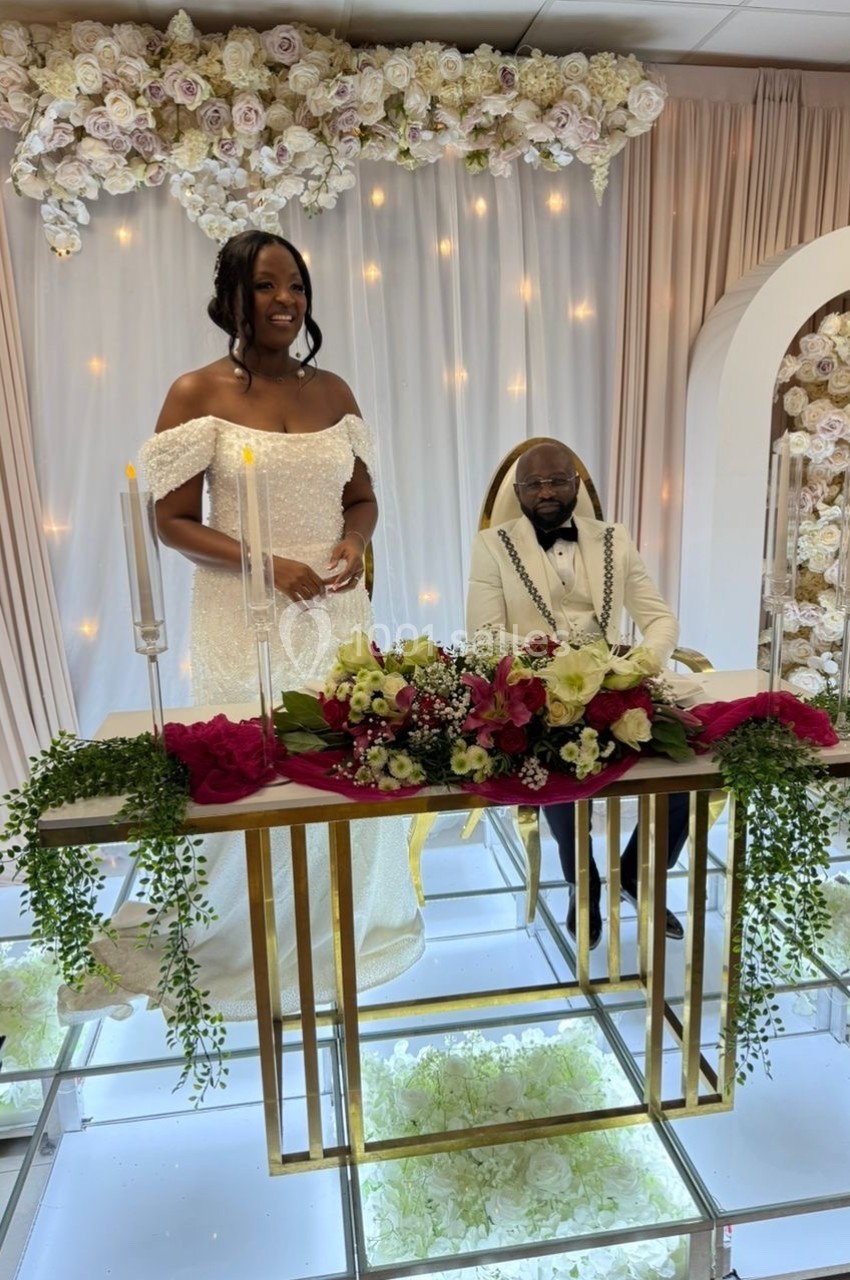 Un couple en tenue de mariage pose devant une table décorée de fleurs et de bougies, avec un fond floral lumineux.