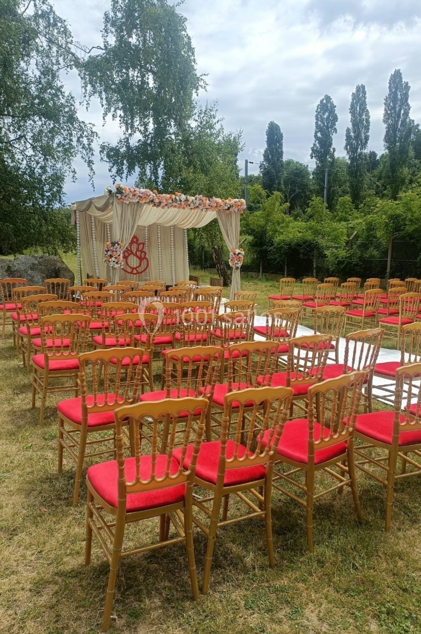 Chaises rouges alignées en extérieur devant une arche décorée pour une cérémonie, entourées de verdure.