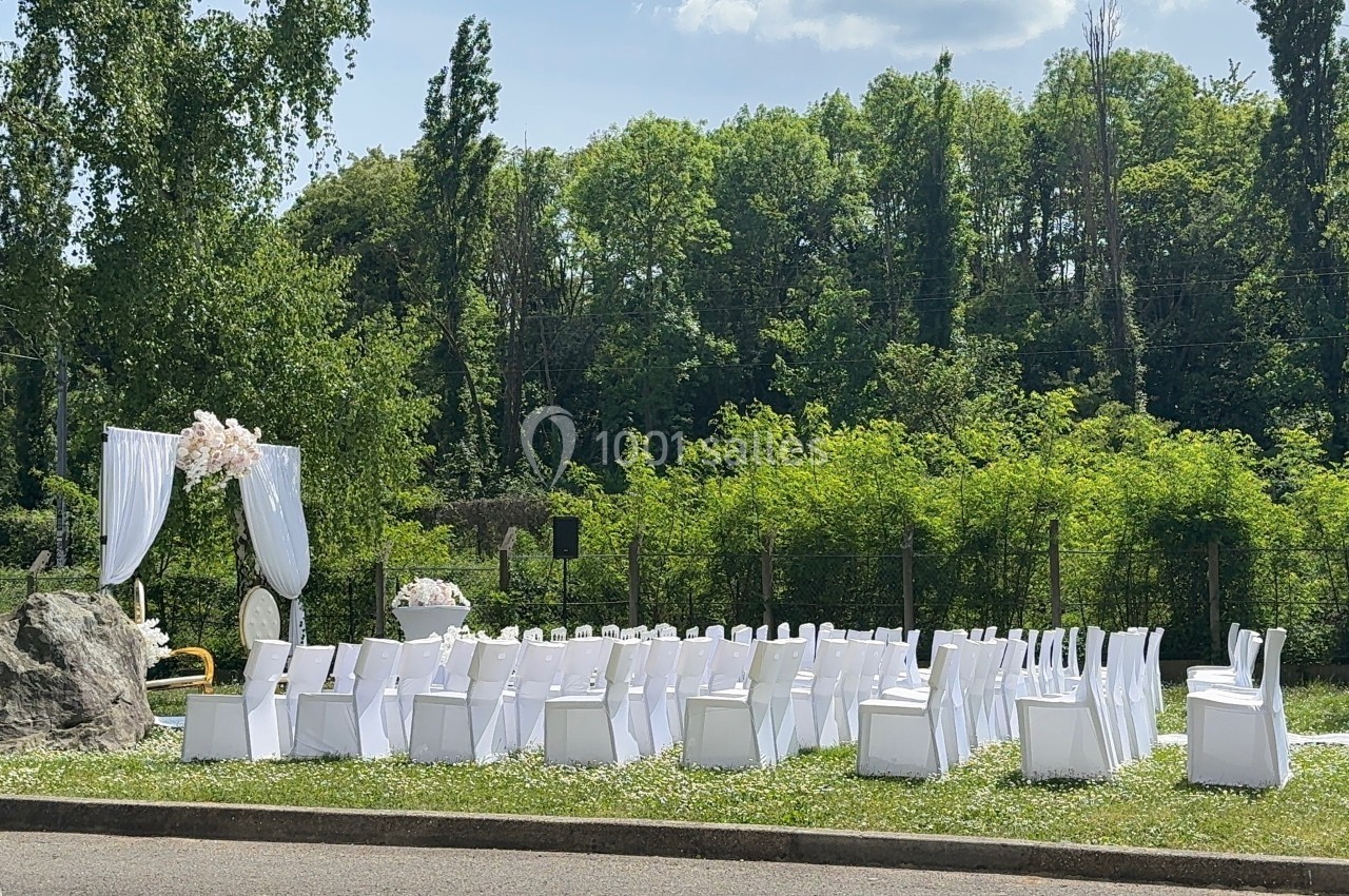 Chaises blanches disposées en extérieur devant une arche décorée de fleurs, entourées de verdure.