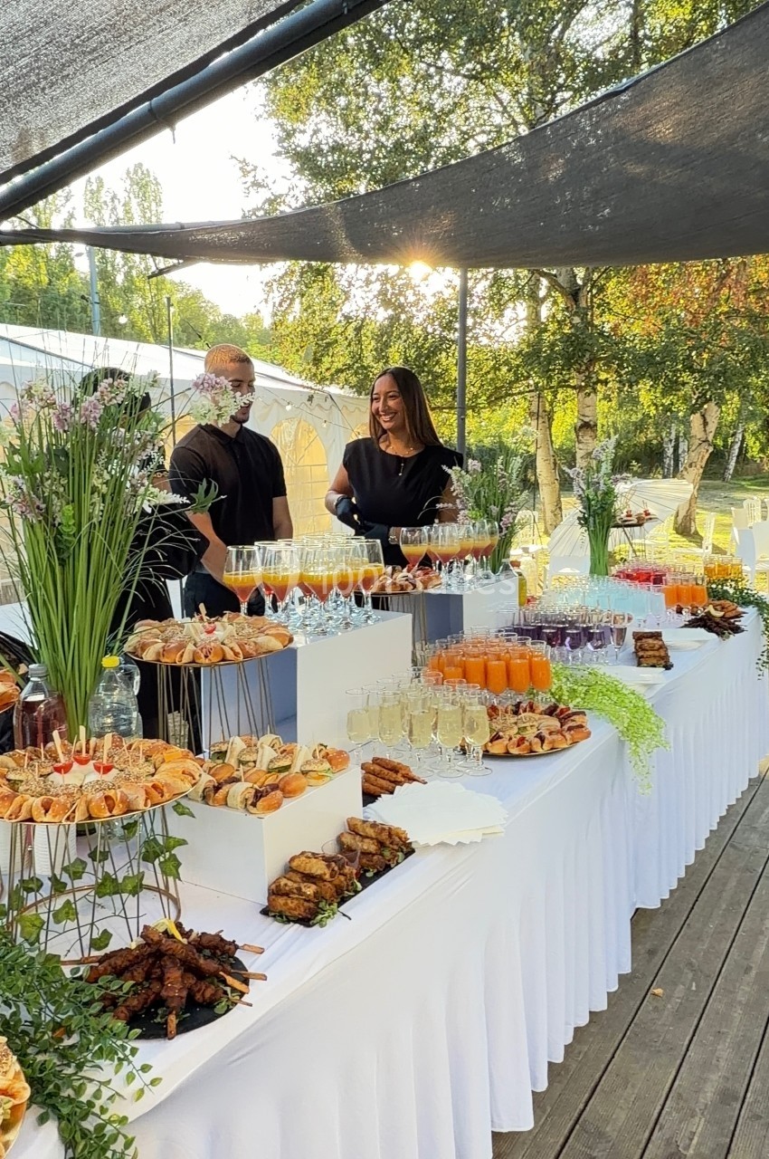 Buffet en extérieur avec divers plats et boissons, deux personnes discutant derrière la table sous un auvent.