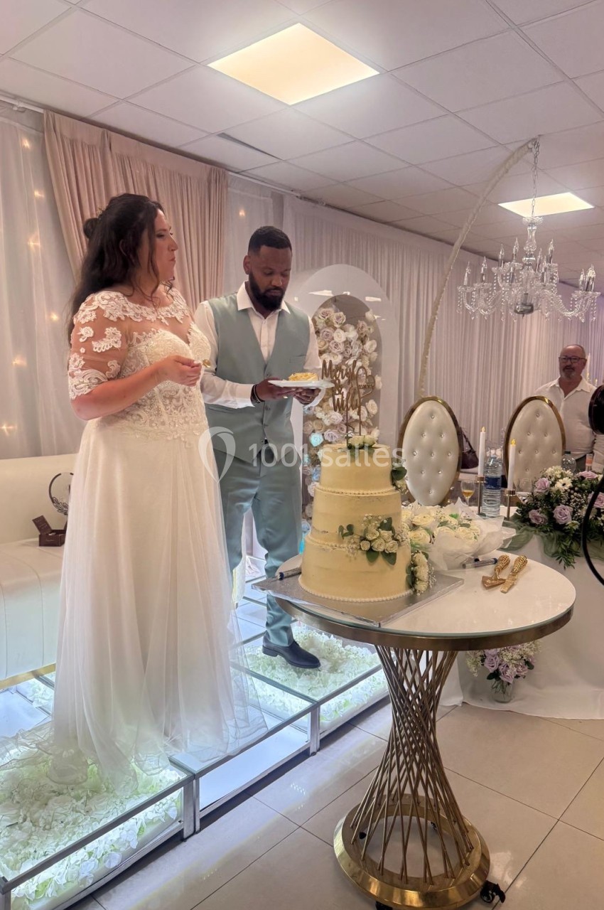 Un couple en tenue de mariage se tient près d'un gâteau décoré, dans une salle ornée de fleurs et lumières.