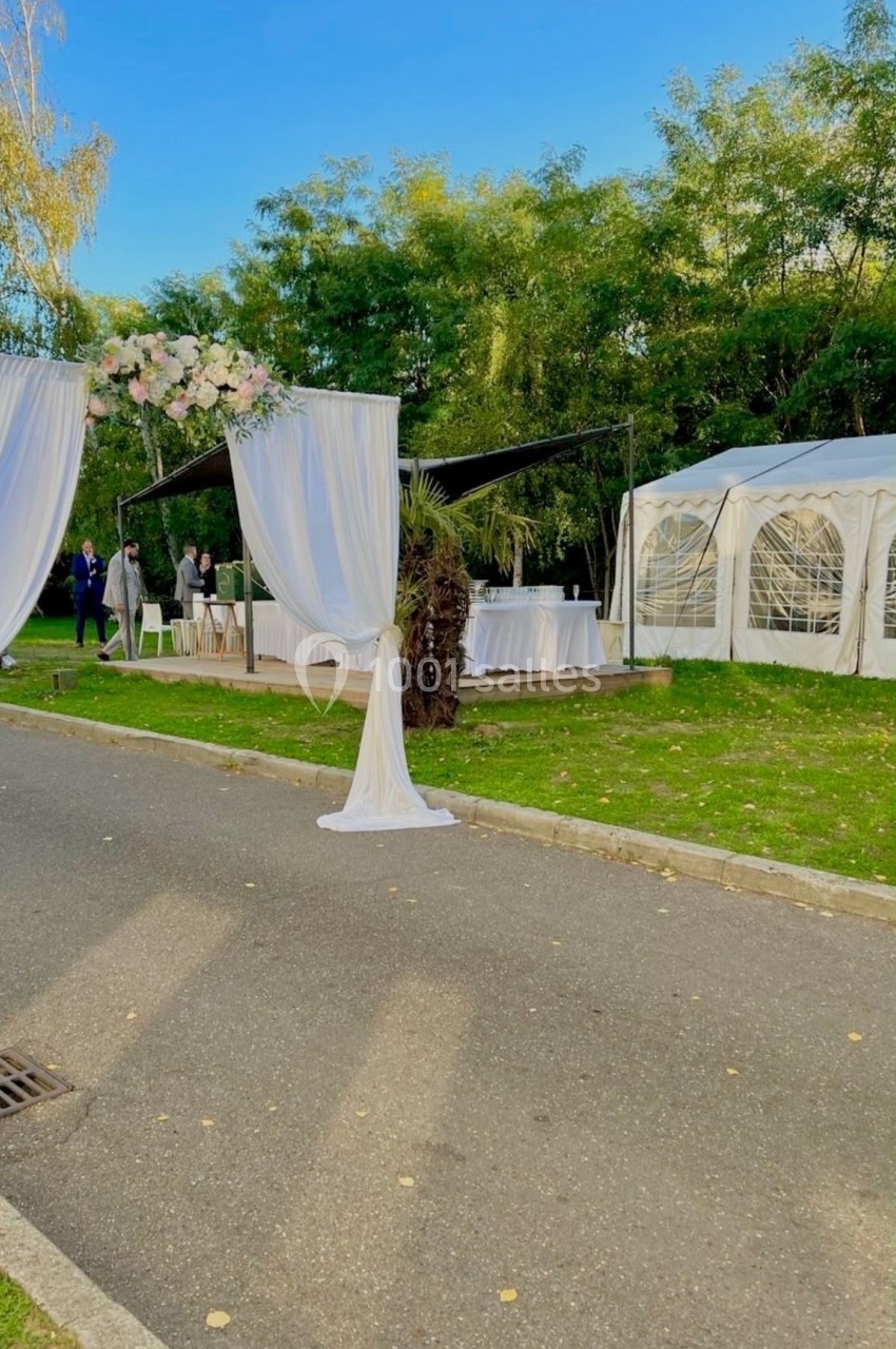 Allée extérieure décorée pour un événement avec des arches fleuries et une tente blanche en arrière-plan.