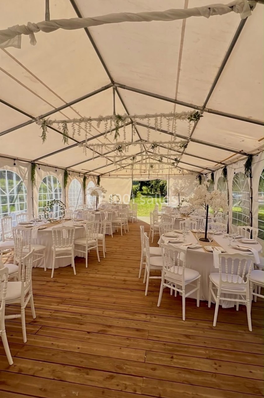Intérieur d'une tente de réception avec tables rondes dressées, chaises blanches et décorations florales suspendues.