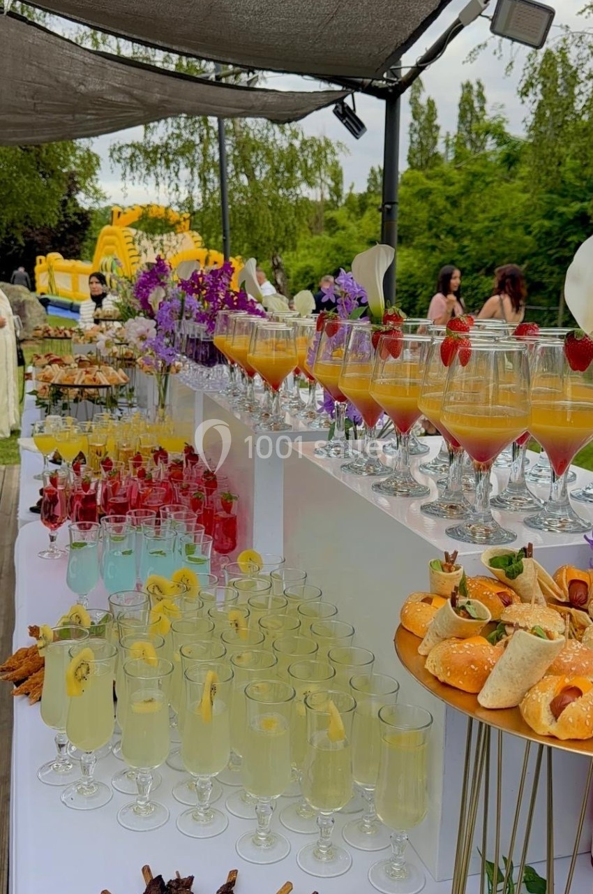 Buffet en extérieur avec cocktails colorés, brochettes, amuse-bouches et décorations florales sous une pergola.