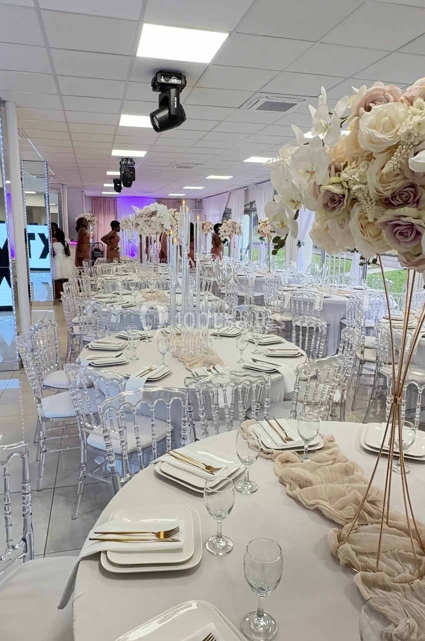 Salle de réception décorée avec des tables rondes, nappes blanches, chaises transparentes et centres de table fleuris.