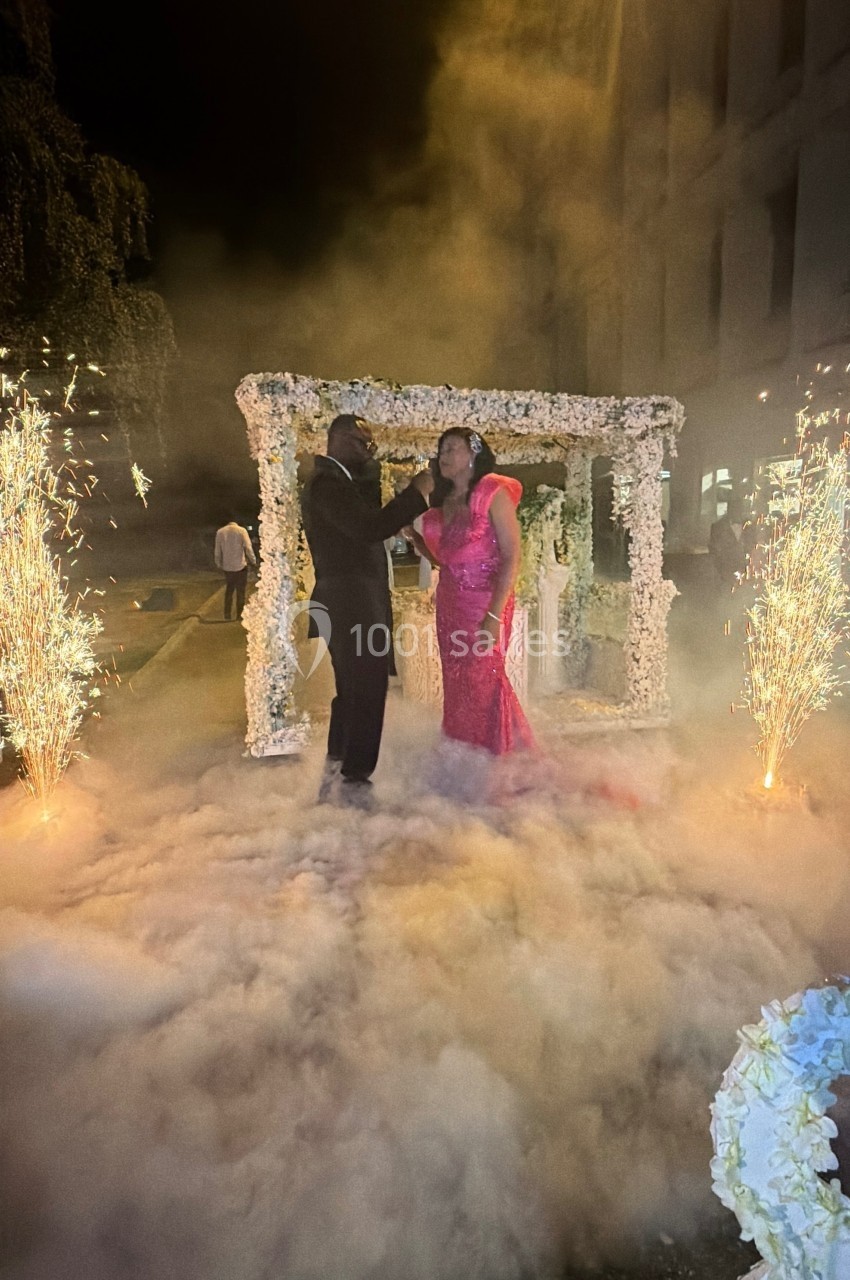 Un couple danse sous une arche fleurie entourée de fumée et de gerbes d'étincelles lors d'une cérémonie nocturne.