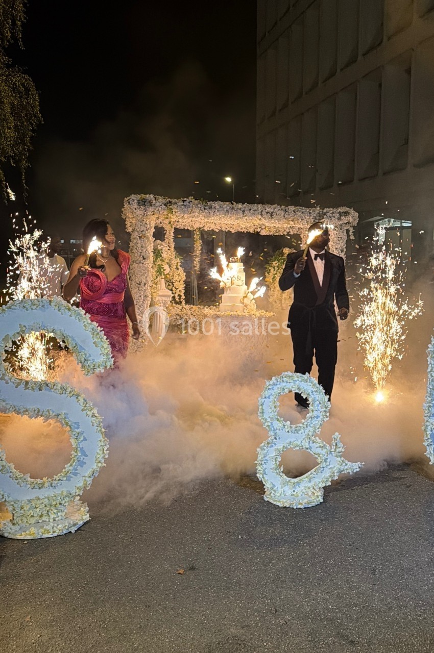 Un couple marche entre des décorations florales et des étincelles, entouré de fumée lors d'une célébration nocturne.