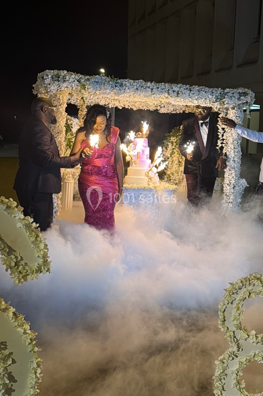 Une femme en robe élégante marche parmi de la fumée artificielle, entourée de décorations florales et d'invités.