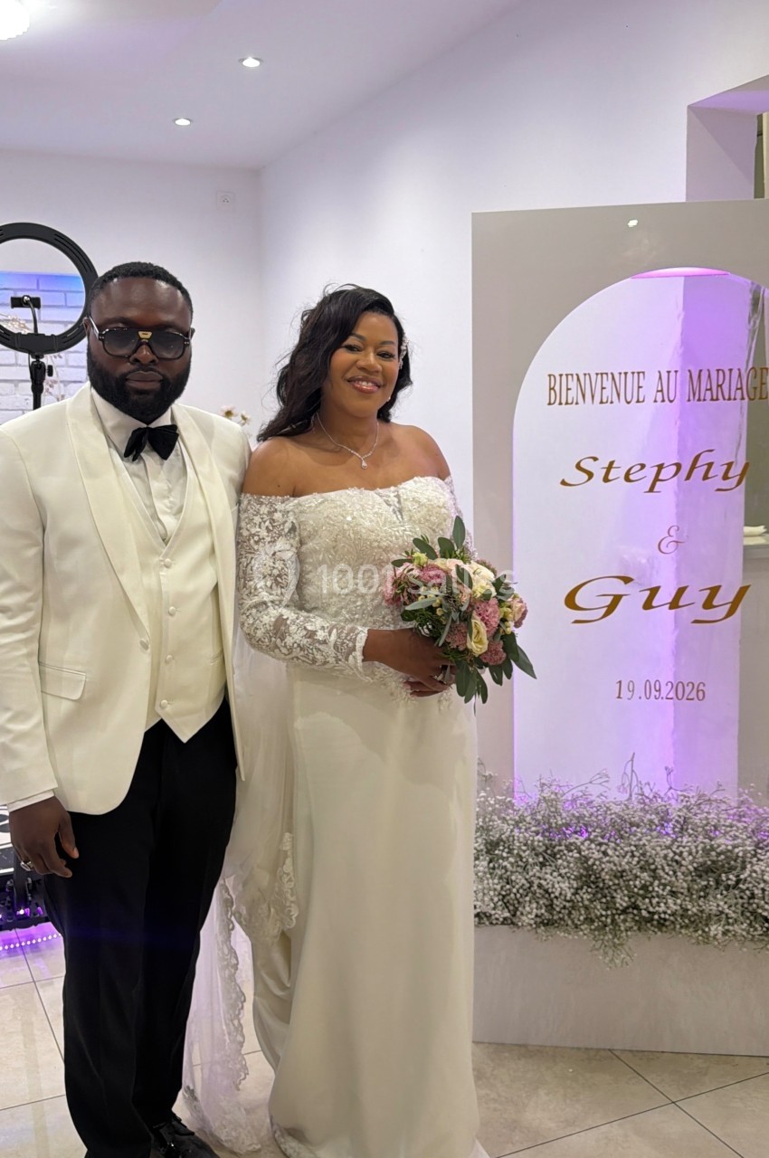 Un couple en tenue de mariage pose devant une décoration florale et un panneau indiquant ’Bienvenue au mariage’.