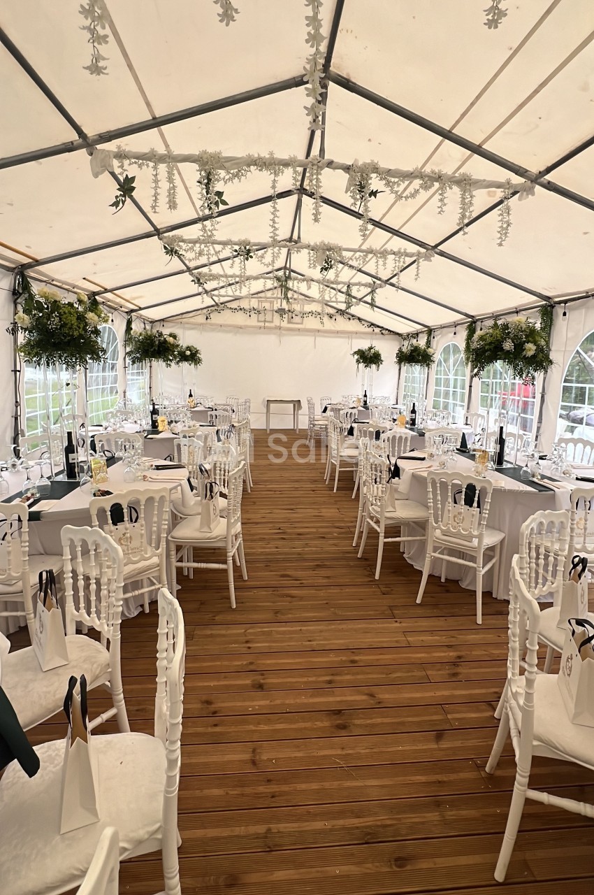 Salle de réception sous une tente blanche, avec tables dressées, chaises blanches et décorations florales suspendues.